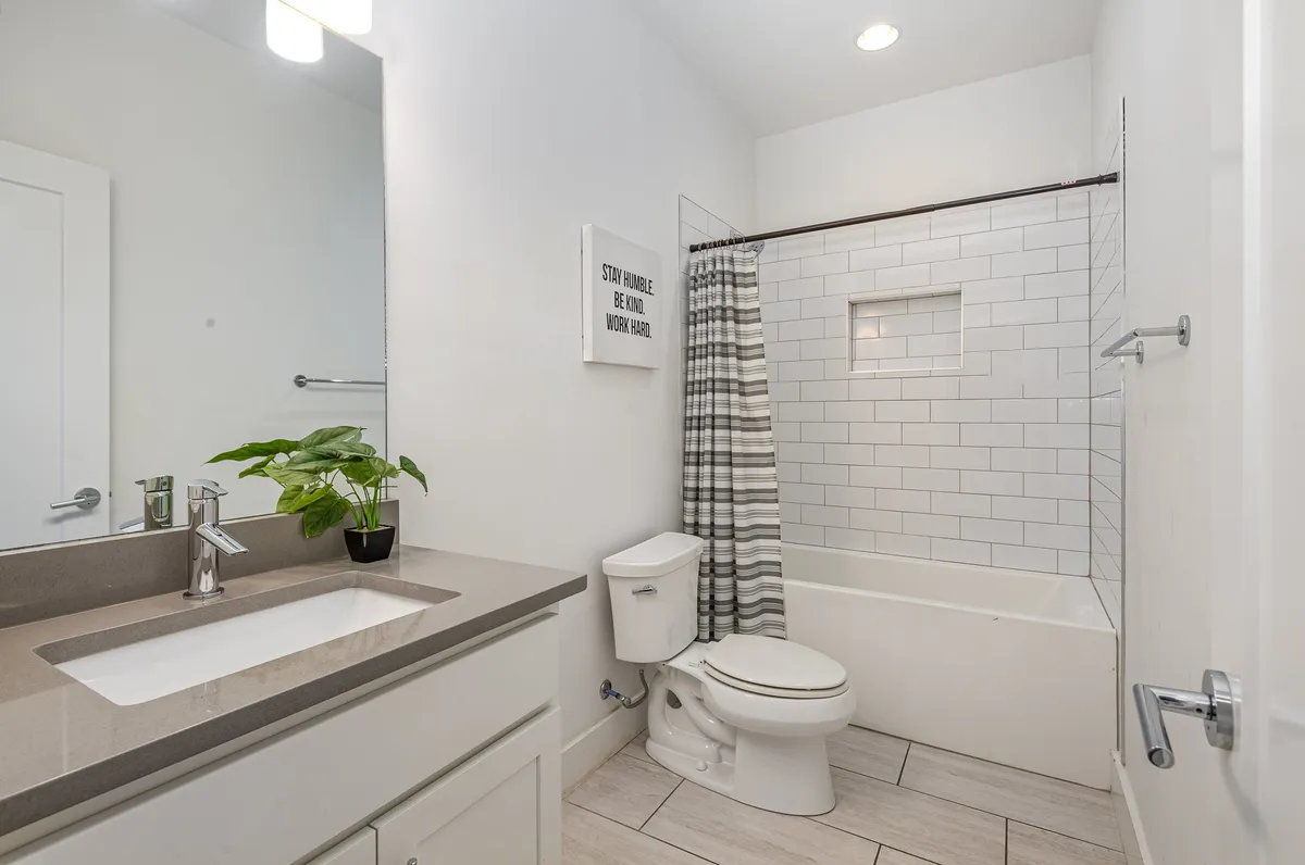 A contemporary secondary bathroom featuring a modern vanity sink, a bathtub with a shower curtain, framed wall art, and a potted plant. The room blends functionality with style, offering both shower and bath options.
