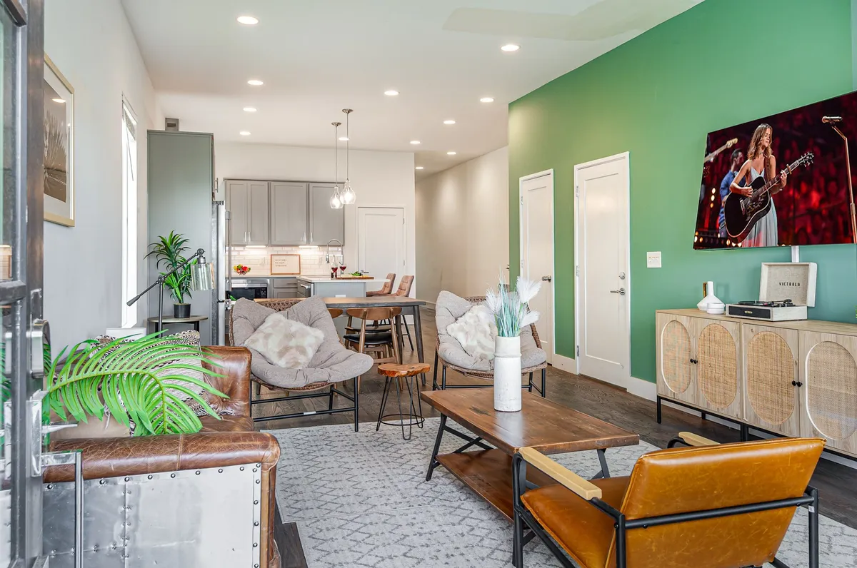 The image captures a stylish, open-concept living area featuring a bold green accent wall, a spacious wooden dining table perfect for group meals, a cozy seating area, and a large TV displaying a music performance. The space is designed for both relaxation and entertainment, with thoughtful decor and a welcoming, casual vibe.