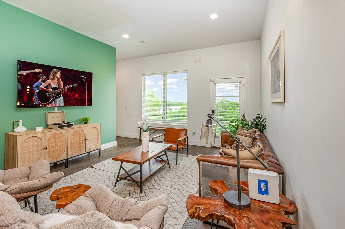 A cozy and contemporary living room with a large television displaying a live performance. The space features a green accent wall, a wooden coffee table, and a comfortable seating area, inviting guests to relax, catch up, or watch TV together.
