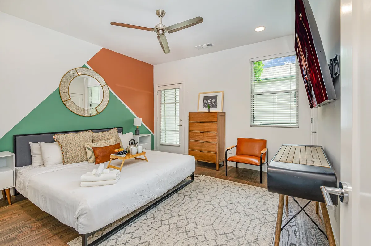 A stylish and cozy bedroom featuring a comfortable bed with an orange accent wall, wooden dresser, armchair, and a wall-mounted mirror. The room is bathed in natural light, offering a tranquil and modern atmosphere perfect for relaxation.